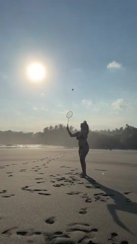 An adult female in a dark bikini is on a sandy beach, silhouetted against a bright, low sun. She holds a badminton racket in her right hand and hits a shuttlecock upwards at the beginning of the clip. Footprints are visible in the wet sand. In the background, there is a body of water and a line of trees, likely palm trees. After hitting the shuttlecock, the woman lowers the racket and walks towards the camera, casting a long shadow on the sand. The sky is light blue with a few clouds.