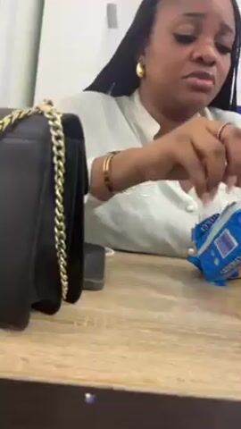 A woman with long, dark braids sits at a light brown wooden table, opening a package of Oreo cookies. She is wearing a light-colored shirt, gold earrings, and a gold bracelet. A black purse with a gold chain sits on the table next to her. A phone is also on the table. The background includes a white wall with a blue sign. The woman appears to be focused on opening the cookie package.