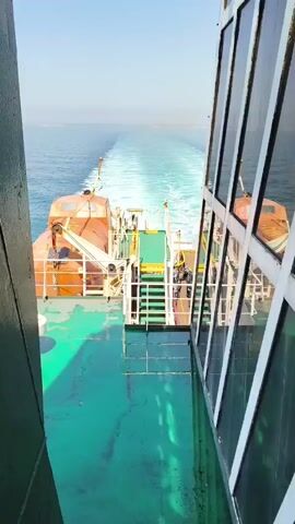 The video shows the wake of a ferry as it travels through the ocean. The ferry has a bright green deck, orange lifeboats, and white railings. There are several people visible on the deck, some of whom appear to be passengers. One person is standing near a motorcycle, suggesting that vehicles are being transported. The sun is shining, indicating it is daytime. The water is calm and a light blue color. The sky is clear and a pale blue. The wake of the ferry creates a foamy white trail behind it, which gradually dissipates into the ocean. The overall scene is one of travel and movement.