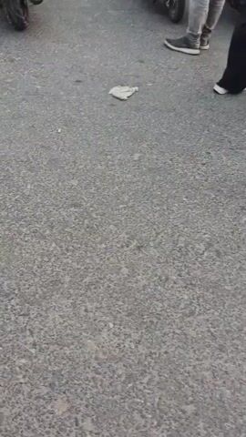 The video begins with a close-up of a mottled grey asphalt surface, with a small, white, unidentifiable object lying on it. The camera then pans up and to the right, revealing the lower body of a person wearing a pink garment and black shoes. Beyond this person, the scene shifts to a street scene with several motorcycles parked. In the background, people are visible near market stalls with colorful awnings. A person in a dark blue hijab and pink garment walks away from the camera. More motorcycles are parked in rows, and several people are walking around them. Further in the background, a BNI bank sign is visible, along with utility poles and buildings. The overall impression is of a busy outdoor market or gathering area.