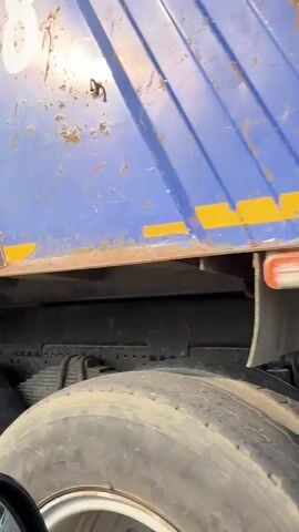 A blue dump truck with white numbers "80" on its side is parked next to an orange truck. The dump truck appears damaged. The camera then pans to a damaged white car with "НОКСТОП" written on its side. Three men are standing near the white car. One man, shirtless and wearing shorts, stands to the left. Another man in a white t-shirt with a red and white logo stands in the center, looking away from the camera. A third man in a bright yellow vest and dark pants is on the right, looking at a phone or tablet. The scene is in a wooded area with dirt and gravel on the ground.
