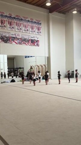 A group of young girls in black leotards are practicing in a gymnastics gym. They are spread out on a large, light-colored mat in front of a wall with large mirrors. Some of the girls are in various dance poses, while others appear to be listening to an instructor who is standing on a raised platform in the background. The gym is well-lit, with fluorescent lights on the ceiling. There are banners hanging on the wall behind the girls, featuring images of athletes and the USA logo. Some gymnastic equipment, including a wooden climbing frame, is visible in the background.