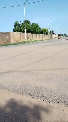 The video shows a sunny outdoor scene on a paved road. In the foreground, the road surface is visible with some texture and debris. A yellow tricycle taxi with two passengers is driving away from the camera on the right side of the road. In the middle ground, a tan brick wall with a sidewalk runs along the right side of the road, with trees and vegetation behind it. Streetlights are visible along the sidewalk. Further down the road, another yellow tricycle taxi is visible in the distance. The sky is clear and blue. The overall impression is a roadside scene in a possibly rural or developing area.