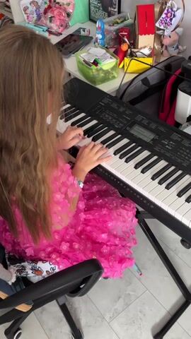 Here's a concise description of the video:

A young girl with long, light brown hair sits in a black office chair, playing an electronic keyboard. She is wearing a bright pink dress adorned with floral appliques. The keyboard is black and sits on a black metal stand. A cluttered desk is visible behind the keyboard, covered with various objects including toys, containers, a phone, and a bag. The floor is light-colored and has a slight pattern. The camera angle is a medium shot from behind the girl, showing her upper body and the keyboard.