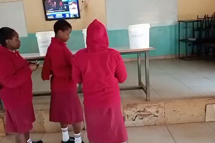 Three young people wearing red uniforms stand in front of a table with four white buckets. The buckets are labeled "ASSISTANT SPEAKER," "ASSISTANT ACADEMIC," "DEPUTY PRESIDENT," and "PRESIDENT." One person is holding a piece of paper. The environment appears to be a classroom with a television screen on the wall displaying a news program. There are chairs stacked in the background.
