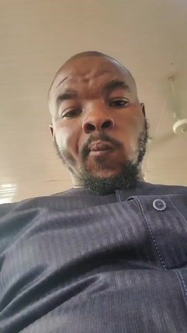 The media file is a close-up, low-angle selfie of a Black man. He is wearing a dark, collared shirt with subtle pinstripes and a button at the neckline. He has a shaved head and a beard. His expression is neutral. The background is a light-colored ceiling with what appears to be a light fixture or fan. The overall lighting suggests an indoor setting during the daytime. The text "00:00" appears in the upper left corner of the video, likely indicating a timestamp. The spoken words are "tomorrow by 4 o'clock." The location context is Numan, Nigeria.