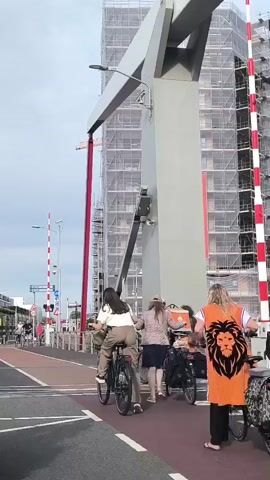 A group of people on bicycles are crossing a street. One person is wearing an orange vest with a lion logo. Another person is carrying a food delivery bag. A building under construction is in the background, covered in scaffolding. There are traffic lights and barriers at the intersection. The weather appears to be overcast.
