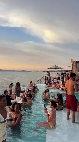 The video shows a poolside party at sunset. The water is calm, and the sky is a mix of orange, yellow, and blue. Several people are in the pool, some are sitting on the edge, and others are standing around. There are lounge chairs and umbrellas on the deck. Some people are drinking, and others are taking pictures. The atmosphere is festive and relaxed.