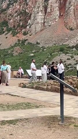 The video captures an outdoor scene with numerous people walking around a large, historic stone building situated against a mountainous backdrop. The building features intricate carvings and external stone stairs. People of various ages, including adults and children, are seen strolling on both dirt and paved paths. A man carries an infant, another man has a child on his shoulders, and a baby is pushed in a stroller. Other individuals are walking, observing the surroundings, or standing in small groups. Some rocks are arranged on the ground, and a small sign is visible.