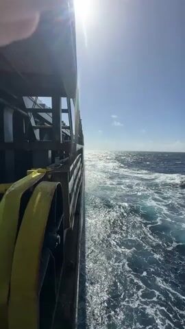 A close-up shot from the side of a boat looking out at the ocean. The sun is shining brightly, casting a glare on the water. The waves are choppy, indicating the boat is moving. Part of the boat's railing is visible in the foreground, along with a yellow strap. The sky is blue with a few scattered clouds.