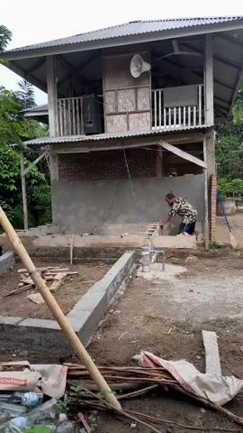 The video captures a scene of construction activity around a two-story wooden building. The building has a metal roof and is partially bricked in on the lower level. A large white speaker is mounted on the exterior of the upper level. A man wearing a patterned shirt and dark pants is actively engaged in building a wooden frame near the building's foundation. The foundation itself is partially constructed with concrete blocks. Various construction materials, including cement bags, wooden planks, and poles, are scattered around the site. The environment appears to be a rural or semi-rural area, with trees and vegetation visible in the background. The ground is a mix of dirt and scattered debris.