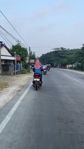 The video shows a rear view of a person riding a motorcycle on a paved road. They are wearing a blue shirt and a pink hijab. Other motorcycles are also visible on the road, some in front and some to the side, indicating a procession or group ride. The environment appears to be a semi-urban or rural area with trees lining the road and buildings on the left side, including a sign that reads "Alfamart." The sky is overcast. The motorcycles are traveling in the same direction. The lighting suggests it is daytime.