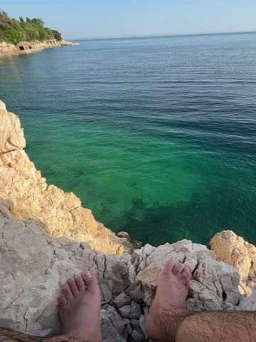 The video opens with a view of bare feet resting on light-colored rocks, overlooking a clear turquoise sea. The camera pans upwards to reveal a broader vista of the coastline. The rocky terrain gives way to a beach nestled at the foot of a cliff. People are visible sunbathing on the sand. The sky is blue with scattered white clouds. The overall setting suggests a tranquil coastal scene on a sunny day.