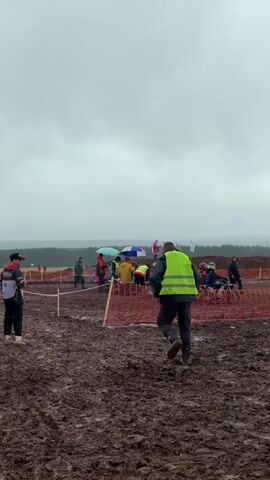This media captures a motocross event in a muddy, outdoor setting under overcast skies, suggesting a cloudy or rainy day. The foreground is dominated by thick, churned mud, indicative of a challenging track. Several people are visible, some wearing high-visibility vests, likely officials or marshals, while others appear to be spectators or participants. A few individuals are standing near what looks like a starting or pit area, with some holding umbrellas, reinforcing the possibility of rain. One person in a bright yellow-green vest is walking away from the camera towards the action. In the background, a line of trees marks the horizon, and a few flags are visible, common at sporting events. The overall atmosphere seems subdued due to the weather. No legible text is visible in the media.