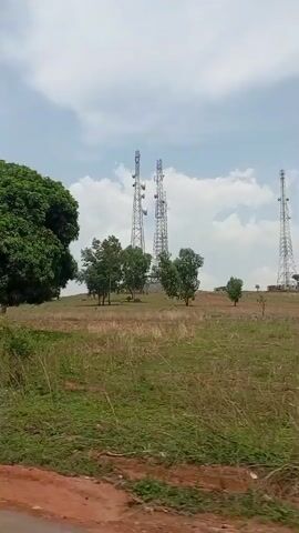 The video displays an expansive, open landscape under a partially cloudy blue sky during daytime. The scene is dominated by a prominent hill in the midground, upon which three tall, metallic lattice-style telecommunications towers are erected, each equipped with multiple antennae.

The foreground features a reddish-brown dirt road or path at the bottom, leading into a field of dry, light-colored grasses and some greener vegetation. Numerous green deciduous trees are scattered across the midground and on the hill itself, providing a natural contrast to the dry terrain. In the far distance, on the right side of the hill, a small, indistinct structure, possibly a building, is visible.

The camera appears to be moving, capturing a panning view of the rural environment. The overall impression is one of a spacious, natural landscape in a rural area like Assob Hausa, Nigeria, where modern communication infrastructure is integrated into the hilly terrain. No people or visible text are present.