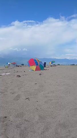 A wide-angle shot captures a sandy beach on a clear, sunny day under a bright blue sky with scattered white clouds. In the foreground, the coarse, light-brown sand stretches out, with a few scattered pieces of debris. In the mid-ground, a colorful beach umbrella, divided into red, yellow, green, and blue sections, stands open, providing shade. A person is visible seated near the umbrella. Further back, a line of parked cars stretches along the horizon, suggesting a gathering or a popular spot. In the far distance, a hazy mountain range is visible under the expansive sky. The overall scene evokes a relaxed, leisurely atmosphere, typical of a day spent at the beach. The time of day appears to be afternoon, given the bright sunlight and the presence of people enjoying the outdoors. There is no visible text in the media file.