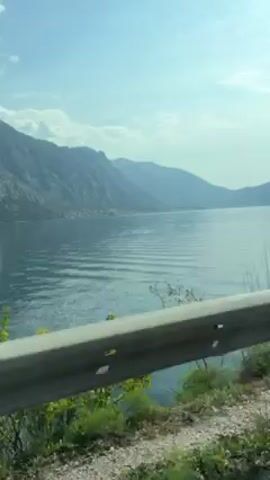 The video shows a drive along a body of water, likely a lake or bay, framed by mountains in the background under a partly cloudy sky. The camera is positioned inside a moving vehicle, with a guardrail visible in the foreground. At one point, the side mirror of the car briefly enters the frame. The overall impression is a scenic drive through a mountainous region.