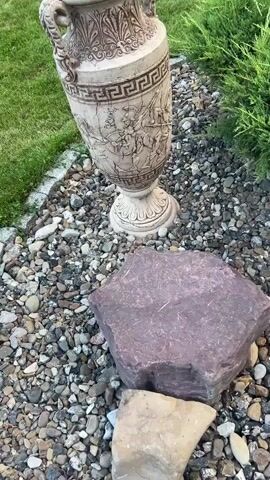 A video begins with a close-up view of a tall, ornate vase resting on a bed of gravel and small rocks. The vase is light in color with a weathered, cracked appearance and features decorative carvings. The top portion of the vase has vertical, leaf-like patterns, followed by a band of Greek key or meander pattern. Below this is a detailed relief depicting a battle scene with figures in ancient armor on horseback and on foot. The vase has two handles that curve upwards and outwards. The background shows green grass and a hint of foliage from a bush or tree on the right side of the frame. A large, reddish-purple rock is positioned in the foreground, partially obscuring the base of the vase. As the video progresses, the camera pans and zooms slightly, offering different perspectives of the vase and its immediate surroundings. In the distance, a white fence with green accents is visible, suggesting an outdoor setting such as a garden or yard. The lighting appears to be natural, possibly indicating daytime. No people are visible in the video. The video focuses exclusively on the decorative vase and its placement in the outdoor environment.