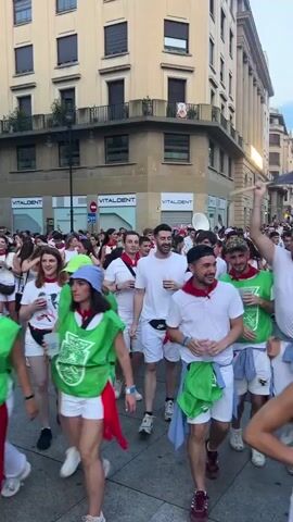 The video shows a lively street scene in Pamplona, Spain, likely during the San Fermín festival. A large crowd of people, mostly dressed in white with red sashes and bandanas, are walking and cheering.

**Subjects:** The primary subjects are the people in the crowd. They are a mix of ages, with many appearing to be young adults. They are dressed in traditional San Fermín attire: white t-shirts and shorts or skirts, complemented by red neckerchiefs and sashes. Some individuals are holding flags, with one prominent flag being waved with yellow and black details on a dark background. Objects include the flags, water bottles, and bags worn by attendees. The scene is a city street lined with buildings, suggesting an urban environment.

**Activity or Event:** The people are participating in a procession or parade, indicated by their movement in a group and the presence of a flag being waved. There is a palpable sense of celebration and excitement.

**Notable Details:** The time of day appears to be late afternoon or early evening, with the sunlight casting shadows. The weather seems pleasant, as people are dressed in light clothing. The setting is clearly identifiable as Pamplona, Spain, by the context of the San Fermín festival and the architectural style of the buildings. The emotions displayed are overwhelmingly positive, with smiles and laughter evident on many faces, conveying joy and festivity.

**Visible Text:** The text "VITAL DENT" is visible on signage on the buildings lining the street. There is also a street sign with a blue circle and a white horizontal bar, common in European signage.