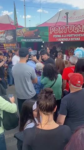 A small crowd of people are gathered in front of a food vendor stand at an outdoor event, likely a festival. The vendor booth has signs advertising "Portugal Pernil a la Portuguesa" and "Estados Unidos." Several people are holding up their phones to take pictures or videos. A man with tattoos is interacting with someone, possibly a customer, while another vendor stands behind the counter. The overall atmosphere seems lively and casual.