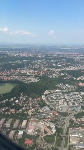 This is a vertical, aerial view of a city on a clear, sunny day. The camera is moving downwards, showing a sprawling urban landscape with a mix of buildings, roads, and green spaces. In the foreground, there are several apartment buildings and residential areas. Further in the distance, the city extends to the horizon, with more dense housing and industrial-looking structures. There is a large, light green, open field bordered by trees. Roads weave through the city, connecting different neighborhoods and appearing as grey ribbons against the urban and natural scenery. The sky is a bright blue with a few wispy white clouds. The viewpoint is from an airplane, as evidenced by the curved edge of an aircraft window visible in the lower left corner of the frame.