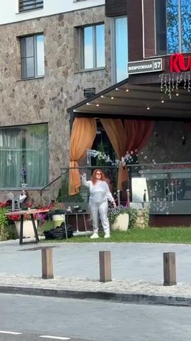 A woman with red hair, wearing a shiny silver jumpsuit and sunglasses, is standing outside a building, making bubbles with a bubble wand. She is in front of a restaurant with orange drapes and a sign that says "Жемчужная 57". There are flowers and plants around her, and a table with a black bag on it to her left. A few people are sitting on a bench in the background. The ground is paved with grey stones.