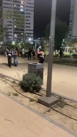 The video shows several young people at night in what appears to be a park or public square. Tall apartment buildings with illuminated windows are visible in the background, suggesting an urban residential setting. Streetlights provide illumination for the area.

In the foreground, two women are actively engaging in a playful water fight or similar activity, spraying each other with what look like water guns or foam blasters. One woman, wearing a black crop top and wide-leg jeans, is holding a white and blue device and spraying the other woman, who is dressed similarly in a black top and dark pants. Another woman slightly further back is also holding a similar device. Other people are scattered in the background, some walking, some standing, near a yellow bench.

The ground consists of paved paths and sections of sand or dirt, with some small bushes and plants visible. A gray trash receptacle is positioned near a small bush in the mid-ground. Partially visible text on the trash receptacle reads "THANK YOU". The overall atmosphere appears casual and lively, with the individuals in the foreground seemingly enjoying themselves.