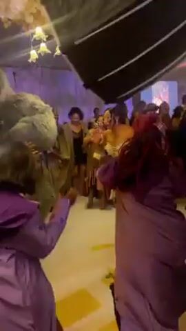 A brightly lit dance floor is packed with people.  Individuals are dressed in colorful, celebratory clothing. The camera moves amidst the dancing crowd, occasionally focusing on specific individuals, but mostly showing the general hustle and bustle of the dance floor.  Decorations, including chandeliers and what appears to be a sculpted green backdrop, are visible.  Some people are filming the event with their phones.