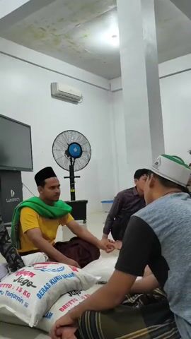 The video shows a group of men sitting on the floor in what appears to be an indoor space. Several large bags of rice are visible in the foreground and around the room. Some of the men are wearing traditional Islamic attire, including peci (caps) and sarongs. One man in the foreground is wearing a grey shirt and a sarong. Another man, wearing a yellow shirt and a green sash, is sitting on the left side of the frame, interacting with the rice bags. In the background, other men are seated, some facing towards the center of the room. An air conditioning unit is mounted on the wall, and a large fan is visible in the background. The overall atmosphere suggests a gathering, possibly for religious or charitable purposes, given the presence of the rice.