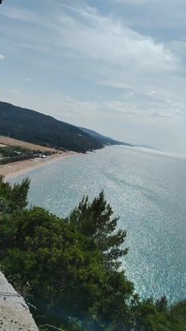 The video captures a scenic coastal view. It begins with a wide shot of the sparkling blue ocean under a partly cloudy sky. The camera then pans right to reveal a long sandy beach on the left, nestled at the base of a verdant hillside. Trees and green foliage are visible along the coast, separating the beach from the water. As the camera continues to pan, it focuses on a historic stone tower perched on a cliff overlooking the sea. The tower is square-shaped with a weathered stone facade, and a set of stairs leads up to its entrance. The blue ocean is visible in the background, stretching out to the horizon.