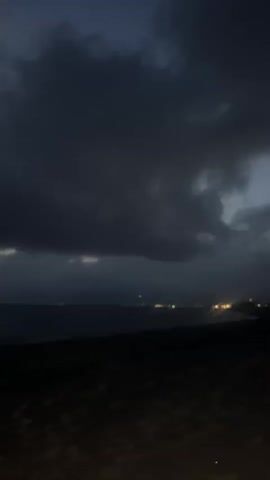 The video shows a dark and cloudy beach at night. The clouds are thick and cover most of the sky, obscuring the moon or stars. In the distance, there are some lights that appear to be from a city or town. The beach itself is dark and sandy, and it's difficult to see any details. The overall impression is of a somewhat bleak and possibly stormy night by the sea.