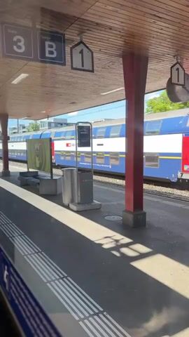 The video captures a view from a train station platform or from inside a train looking out. In the initial frames, a blue and white single-deck train passes from right to left on a track. Behind it, a platform with a red vertical pillar, two grey trash bins, and an advertisement board is visible. Overhead, a wooden ceiling structure features hanging signs with numbers "1" and "2" and letters "B". A digital information display shows "S5 15:45", "Pfäffikon SZ", and "Wetzikon". A clock is also mounted on the ceiling structure. After the first train passes, a stationary blue and white double-decker train, bearing the "SBB CFF FFS" logo, is fully revealed at the platform. Sunlight creates long shadows on the platform surface. Trees and buildings are visible in the background.