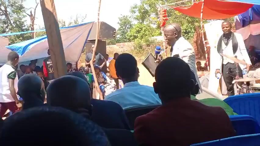 Here's a descriptive summary of the video:

The video shows a gathering of people outdoors, seemingly at an event under makeshift shelters made of tarps and fabric. An older man in a light-colored suit is speaking into a microphone while holding a tablet or book. Another man in a white suit stands behind him. The crowd faces the speaker, and some children are visible in the foreground under one of the shelters. Some people are seated in chairs, and others are standing. Trees and a few buildings are visible in the background. The scene appears to be captured with a handheld camera, from the perspective of someone in the audience.
