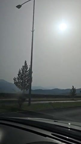 The video shows a view from inside a vehicle looking out at a landscape. The sun is bright and high in the sky, casting a glare. In the middle ground, there is a road with a grassy median and a few trees. In the background, there are mountains. The windshield wipers are visible at the bottom of the frame. The scene appears to be a highway or road passing through a rural or semi-rural area.