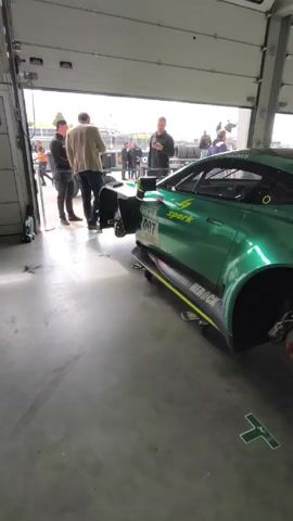 The camera pans around a green race car inside of a garage. The car has no tires on and there are several people talking in the background, outside the garage. The garage is filled with race tires and other equipment.  The car is being worked on or is in the process of being worked on.