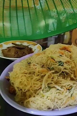 A close-up shot showcases a plate piled high with cooked vermicelli noodles, garnished with shreds of carrot and some green vegetable, possibly spinach or spring onion. The noodles have a pale yellow hue. Partially visible behind the plate is a dish containing a dark-colored food item, perhaps meat or beans. A green plastic food cover hovers above the noodles, out of focus in the background. The scene appears to be indoors. The plate of noodles and the accompanying dish rest on a shiny surface, possibly a table.