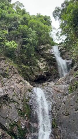 The video shows a multi-tiered waterfall cascading down a rocky, overgrown mountainside. Lush green trees and foliage frame the scene, with the water flowing from a higher elevation to a pool at the bottom. The rocks are varied in texture and color, with some appearing moss-covered. The sky is overcast, suggesting a natural, outdoor environment. There are no people or man-made objects visible.