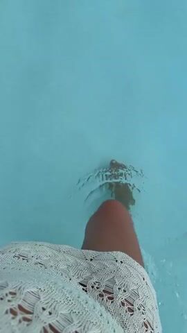 A person's legs and feet are submerged in a pool of clear blue water. The camera is positioned above the water, looking down at the person's legs. The person is wearing a white lace cover-up over what appears to be a bathing suit. The water is disturbed by movement, creating ripples and splashes. The person's toenails are painted white.