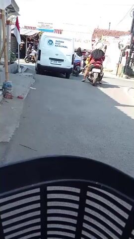 A white van with "BADAN GIZI NASIONAL" written on its back is driving down a street. Multiple people on motorcycles are following behind the van. The street appears to be in a village or a rural area, with buildings on either side of the road. The camera is mounted low, looking forward from what appears to be the perspective of someone riding on a motorcycle. The overall scene is one of traffic on a sunny day.