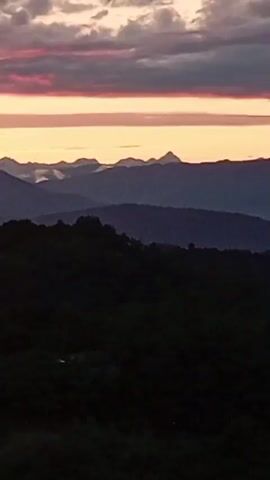 The video shows a slow zoom out of a mountainous landscape at sunset. The sky is filled with dramatic clouds painted in shades of pink, purple, and orange. Several layers of dark, silhouetted mountains recede into the distance, with a lighter blue haze softening their contours. The foreground is dominated by a dark, forested area, obscuring any details within it. The overall impression is one of a peaceful, natural scene as day transitions into night.