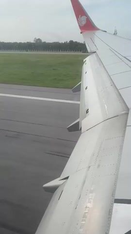 The video shows the view from the window of an airplane during takeoff or landing. The plane's wing is visible in the foreground, with flaps extended. The runway and surrounding grassy area are visible beyond the wing. In the distance, a tower and trees can be seen. The plane appears to be moving on the ground or ascending rapidly. The overall lighting suggests it is daytime.