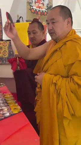 Two men wearing monastic robes are performing what appears to be a Buddhist ritual. The man in the bright yellow robe is holding a peacock feather in one hand and a bell wrapped in red cloth in the other. He is gesturing over a table lined with small bowls filled with liquid. The other man, wearing a maroon robe, is seated and watches the actions of the first man. Behind them is a yellow cloth with a religious image on it. The room appears to be a temporary structure, possibly a tent.