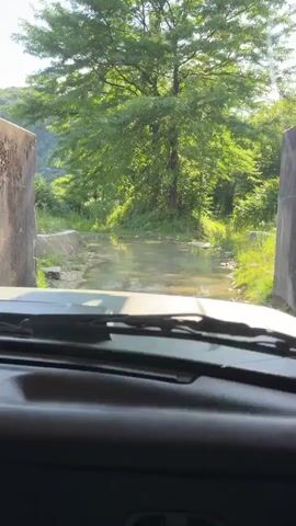The video is filmed from the interior of a vehicle, looking forward through the windshield. The vehicle is driving on a dirt road that has several puddles of water. Trees and lush green vegetation line the sides of the road and the surrounding hills. The sun is shining, casting dappled light through the trees. The driver's side windshield wipers are visible in the foreground.