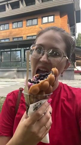 A woman with glasses and a red shirt is eating a bubble waffle ice cream cone outside a wooden building. She is holding the cone in her right hand and has her mouth open, taking a bite. The ice cream has pink swirls and is topped with blueberries and chocolate sauce. Her left hand is holding the cup the cone is in. The building in the background is made of wood and has multiple stories with windows. The sky is cloudy.