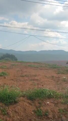 The video shows a rural landscape with rolling hills in the background, transitioning to a more developed area. It starts with a view of a field with red soil and scattered greenery, under a partly cloudy sky with power lines overhead. The camera then pans to the right, revealing a structure, possibly a workshop or garage, with equipment and objects visible inside. Continuing the pan, the camera captures a scene with a sign, some trees, and a small building in the background. The video concludes with a view of a property featuring two parked trucks and a house or building set against the backdrop of distant hills. The ground is muddy, suggesting recent rain.