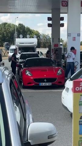 A red Ferrari is parked at a gas station. A person in an orange reflective vest is walking away from the car. Another person is standing by the pump, appearing to refuel the car. A white camper van is parked behind the Ferrari. Several other vehicles are visible in the background. The environment appears to be an outdoor gas station with a canopy overhead.