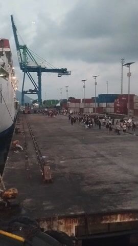 The video shows a port scene from a low angle, with a ship in the foreground. A large crane with blue and green parts is visible. In the background, there are stacks of shipping containers and several people standing on the dock. The sky is overcast. The camera appears to be tilted, making the scene appear vertical.
