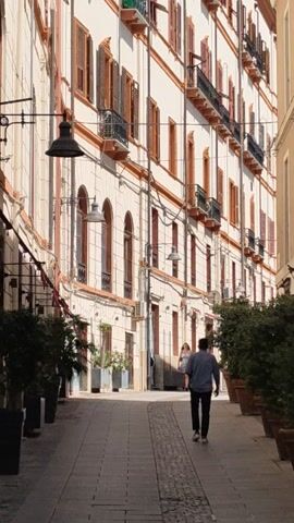 A pedestrian street is framed by buildings on either side. The buildings are a pale color with rows of windows and architectural details. People are walking along the street. Trees and street lamps are present. The video is shot at an angle, making the scene appear sideways.