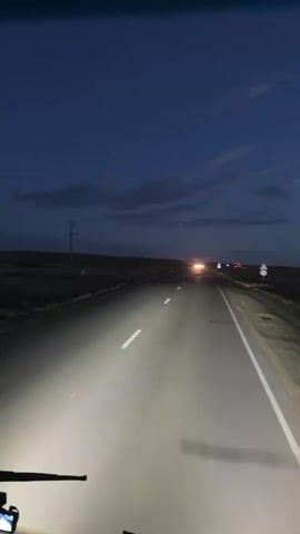 This media file captures a first-person perspective of driving on a highway at dusk. The scene is set on a paved road stretching into the distance, with a dark, twilight sky overhead, showing wisps of clouds. Utility poles are visible along the roadside, suggesting a rural or semi-rural setting. The headlights of the vehicle are on, illuminating the road ahead. In the distance, other vehicles are visible, their headlights creating bright points of light. The overall mood is serene and perhaps a bit lonely, typical of a solitary drive as daylight fades. There is no visible text in the media file. The location context is Baiqadam, Kazakhstan.