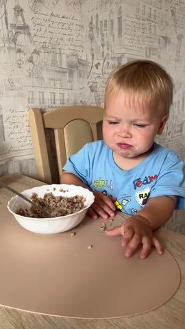 A young child with blonde hair and blue eyes, wearing a blue t-shirt, sits at a table. The child is positioned in front of a white bowl filled with a grainy, brown food, and a silver spoon. The child's left hand rests on the table, and their right hand holds the spoon, which is inside the bowl. The child is looking down at the bowl with a concerned expression. The table is covered with a beige placemat, on which some of the food has spilled. In the background, a wall is decorated with a patterned wallpaper featuring images of Parisian landmarks. The child is seated in a wooden high chair.